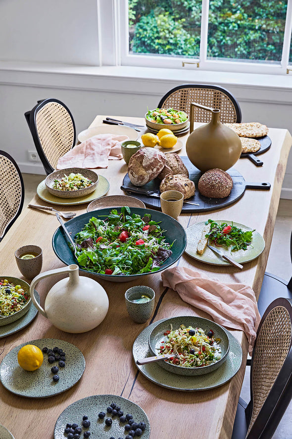 A tablescape viewed from above. Plates of food sit around the edge of the table, while a big bowl of salad sits in the middle. Two round ceramic jars, one mustard and one white, sit at either end of the table.