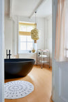 View into a bathroom, a black bath sits in the centre of the room. On the window ledge are two ceramic vases. A large round mustard coloured one and a smaller grey one.