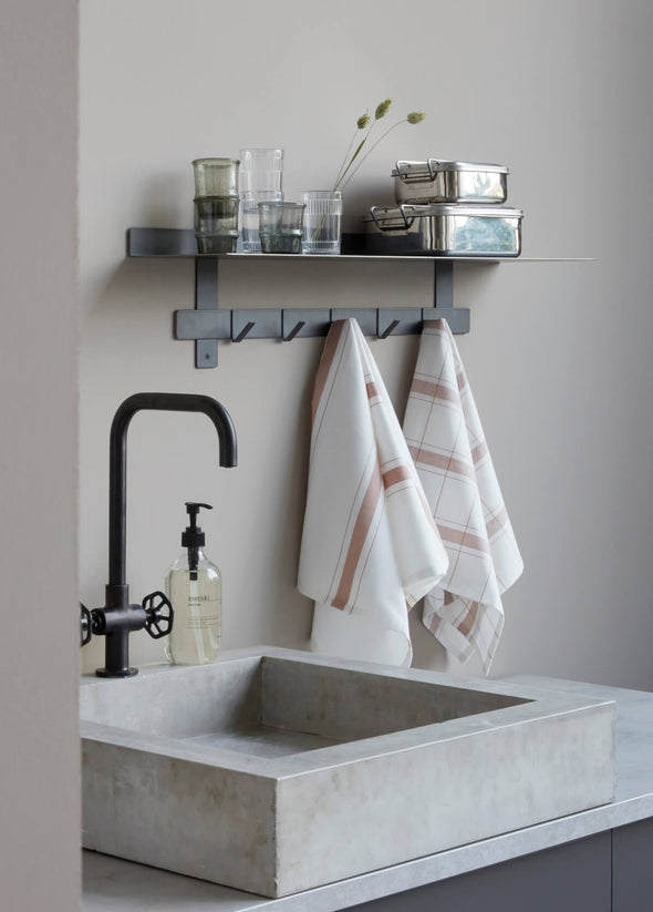 A concrete square sink and black tap. On the wall behind is a metal shelf with hooks below. On the shelf are assorted drinking glasses and two metal storage tins. Hanging from the hooks are two kitchen towels. They are white with a nougat stripe and check design.