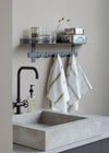 A concrete square sink and black tap. On the wall behind is a metal shelf with hooks below. On the shelf are assorted drinking glasses and two metal storage tins. Hanging from the hooks are two kitchen towels. They are white with an olive green stripe and check design.