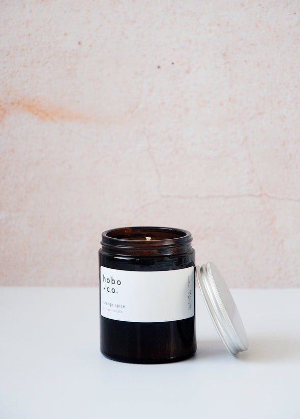Scented candle in a brown apothecary inspired jar which is open with an aluminium lid leaning against the side, made by HoBo + Co Candles using only natural soy wax. The label shows the scent as being Orange Spice. The jar has a capacity of one hundred and eighty millilitres and is eight centimetres tall and six point five centimetres wide.