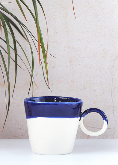 Porcelain mug, handmade by Jade Gallup, with a dark blue glaze over the top third and white glaze on the rest with a perfectly round handle on the side. Eight centimetres tall and twelve centimetres wide including the handle.