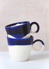 Two porcelain mugs, handmade by Jade Gallup, stacked on top of each other. Each has a dark blue glaze over the top third and white glaze on the rest with a perfectly round handle on the side. Each mug is eight centimetres tall and twelve centimetres wide including the handle.