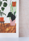 Close up corner of an art print by Rachel Victoria Hillis. Depicting a collection of potted green houseplants, some on the floor, some on stools and some hanging. The pots are pink, terracotta and brown, on a white background and wooden planked floor.