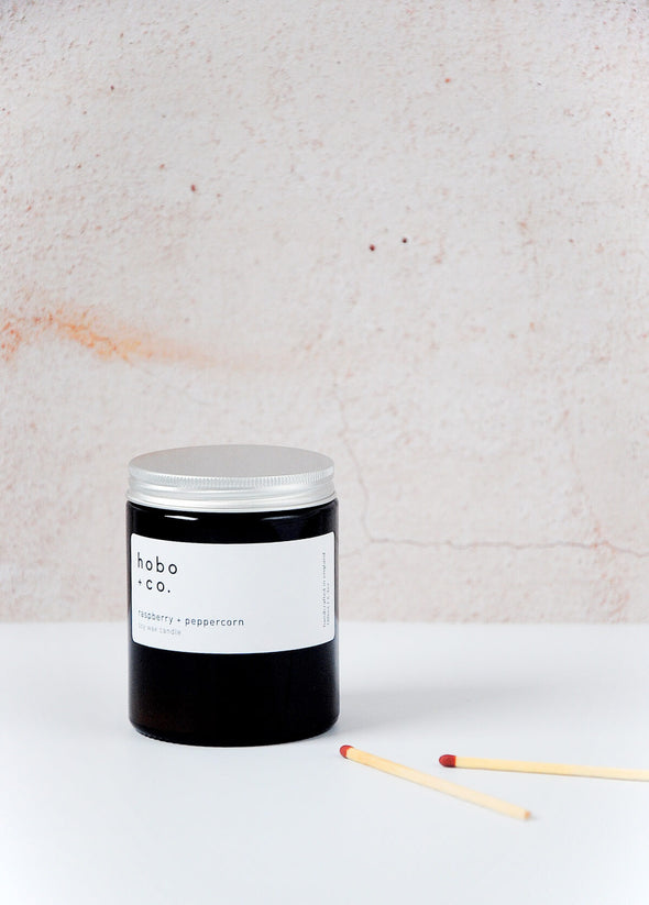 Scented candle in a brown apothecary inspired jar with an aluminium lid, made by HoBo + Co Candles using only natural soy wax. The label shows the scent as being Raspberry and Peppercorn. The jar has a capacity of one hundred and eighty millilitres and is eight centimetres tall and six point five centimetres wide.