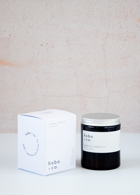  Scented candle in a brown apothecary inspired jar with an aluminium lid, made by HoBo + Co Candles using only natural soy wax. Next to the candle is the white packaging which consists of a simple white cardboard box with the product details and branding printed in black. The label shows the scent as being Raspberry and Peppercorn. The jar has a capacity of one hundred and eighty millilitres and is eight centimetres tall and six point five centimetres wide.