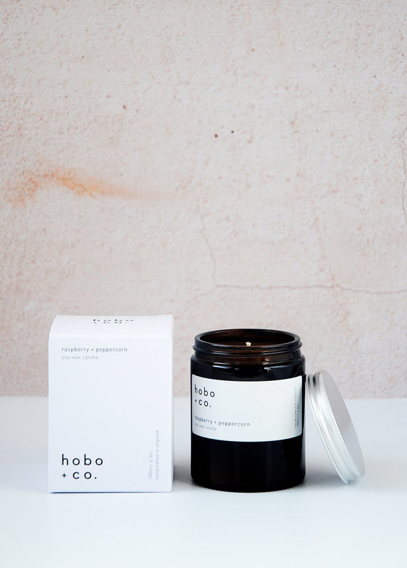 Scented candle in a brown apothecary inspired jar which is open with an aluminium lid leaning against its side, made by HoBo + Co Candles using only natural soy wax. Next to the candle is the white packaging which consists of a simple white cardboard box with the product details and branding printed in black. The label shows the scent as being Raspberry and Peppercorn. The jar has a capacity of one hundred and eighty millilitres and is eight centimetres tall and six point five centimetres wide.