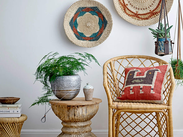Interiors image featuring a side table with a vase and plant pot on top, alongside a wicker chair with a red cushion resting on it.