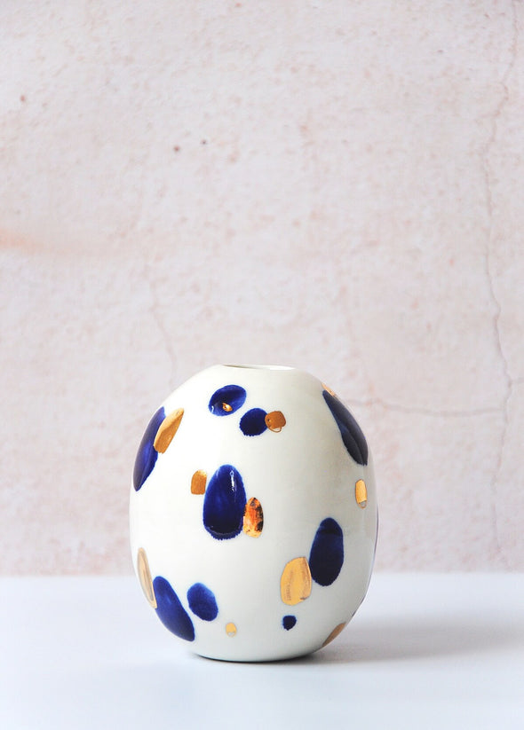 Oval shaped porcelain bud vase, handmade by Jade Gallup. Featuring a white glaze base and covered with splodges of deep blue and real gold lustre. Can be used to display one or two stems, or alone as a decorative piece. Approximately eleven centimetres tall.