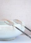 A pair of salad servers made from stainless steel. One has the shape of a spoon and the other the shape of a fork. The handles are long and thin with a darker, rustic finish. Each is thirty centimetres long.