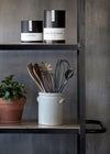 Salad servers in a white ceramic utensil jar along with a selection of other utensils. The jar sits on a metal shelving unit next to a terracotta plant pot containing a green plant. On the shelf above is a box of tea and a box of coffee. The background wall is a medium grey colour.