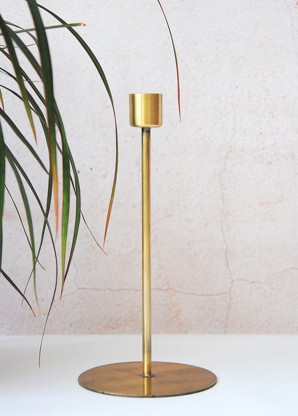 Individual metal candle stand, featuring a wide disc as a stand at the bottom and tall thin body. Made of iron with a brass finish. Suitable for candles measuring four centimetres in diameter. Twenty centimetres tall, the bottom disc is nine point five centimetres wide.