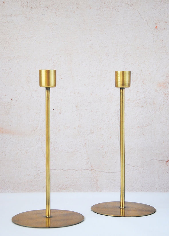 Two metal candle stands, featuring a wide disc as a stand at the bottom and tall thin body. Made of iron with a brass finish. Suitable for candles measuring four centimetres in diameter. Twenty centimetres tall, the bottom disc is nine point five centimetres wide.