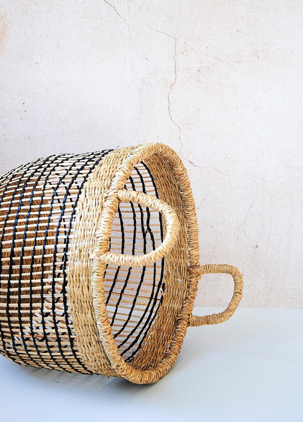 Small open weave seagrass basket, lying on its side, in natural and black with handles. Height twenty six centimetres, width thirty one centimetres.
