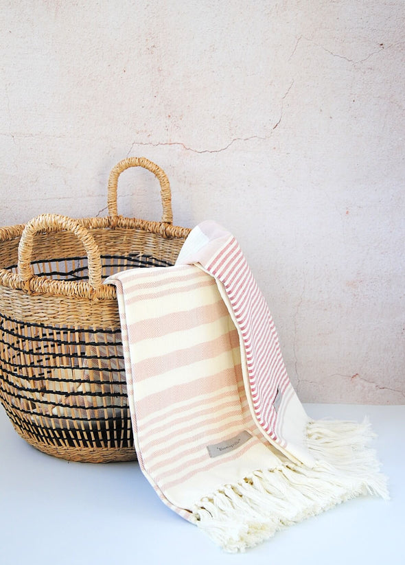 Small open weave seagrass basket in natural and black with handles. A set of two cream and rose towels draped over the side.