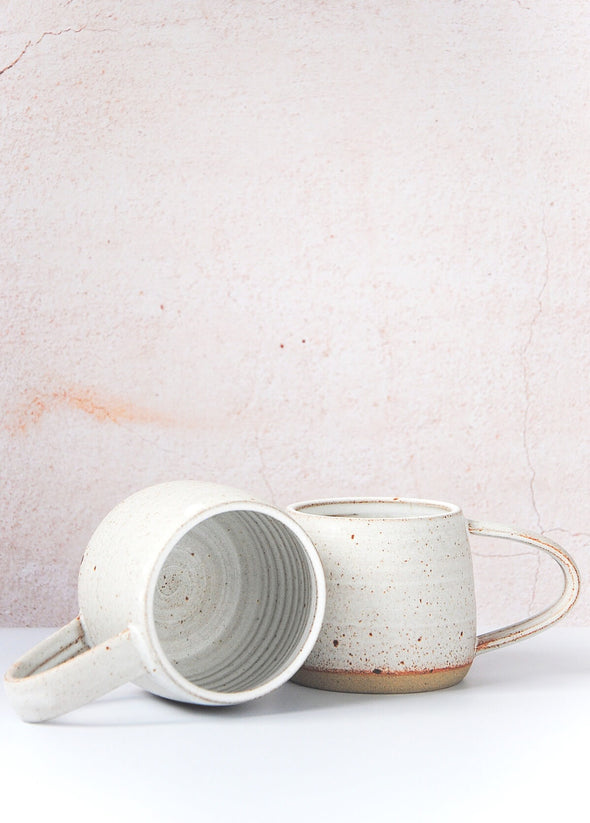 Two stoneware mugs, one upright and one on it's side. Handmade by JG Pottery, with a white glaze and rustic finish.