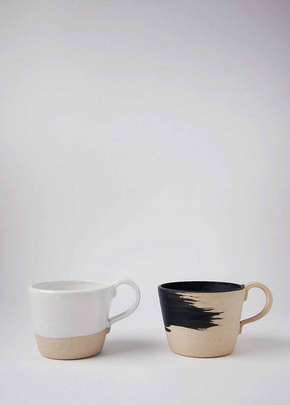 Two stoneware mugs sitting side by side. One has a raw finish and a matt black brush stroke design on the exterior. The interior of the mug is painted in a matt black glaze. The other is finished in a glossy white glaze.