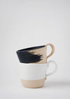 Two stoneware mugs in a stack. One has a raw finish and a matt black brush stroke design on the exterior. The interior of the mug is painted in a matt black glaze. The other is finished in a glossy white glaze.