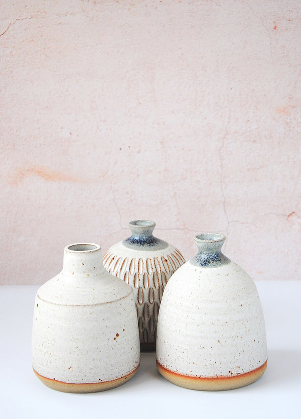 Group of three stoneware bud vases, handmade by JG Pottery. One bottle vase featuring a white glaze, one dome shaped vase featuring a white glaze on its body and a blue over dip glaze on its neck and one dome shaped vase with decorative carving featuring a white glaze on its body and blue over dip glaze on its neck.