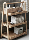 A rattan shelving unit consisting of three shelves. The top shelf displays a basket containing toiletries, the middle shelf houses a metal bowl and a pile of folded hand towels in a cream and grey stripe design. The bottom shelf contains a grey storage box.