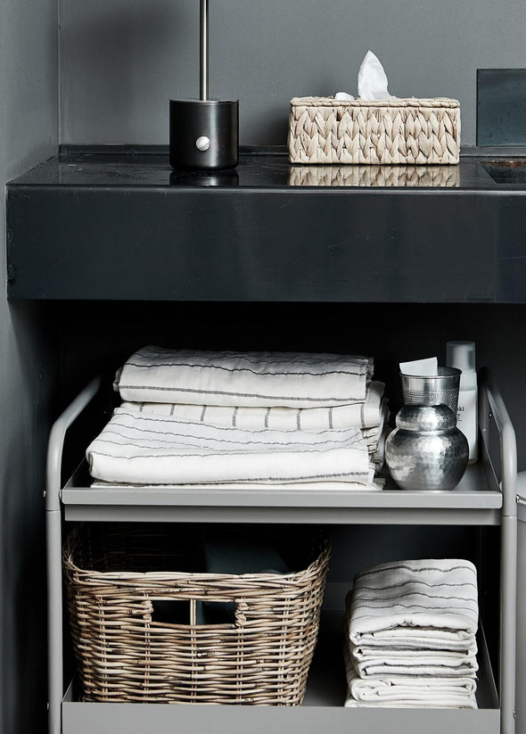 A black sideboard under which there is grey metal storage trolley. On the trolley are two piles of folded towels in a cream and grey stripe design, alongside a basket and metal pots.