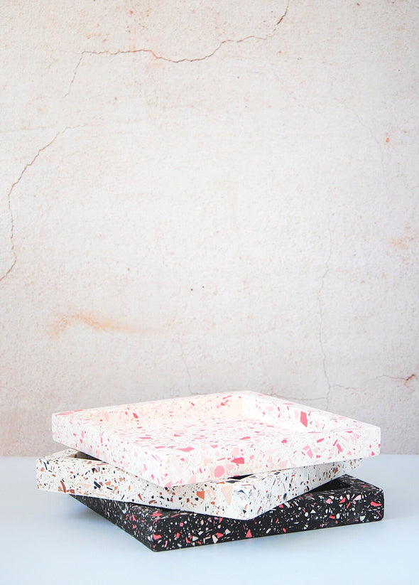 A stack of three terrazzo square trays handmade by Katie Gillies using jesmonite. The bottom tray has a black base with red, pink and white chips. The middle tray has a white base with brown, pink and black chips. And the top tray has a white base with pink and red chips. Each tray is twenty centimetres by twenty centimetres and two centimetres thick.