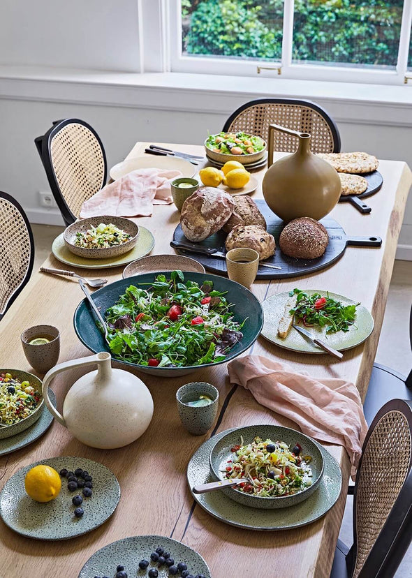 A tablescape viewed from above. Plates of food sit around the edge of the table, while a big bowl of salad sits in the middle. Two round ceramic jars, one mustard and one white, sit at either end of the table.