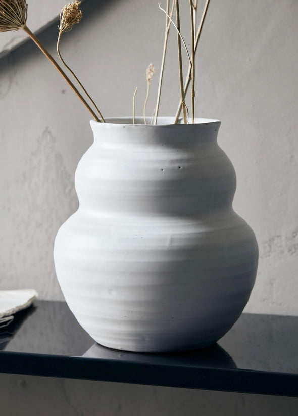Ceramic vase with an organic rounded shape and simple white glaze. It is displayed on a shelf holding a few stems of dried flowers. The vase is 19cm tall and 17cm wide.